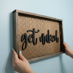 Framed wooden sign that says "get naked" in cut wood with a herringbone pattern engraved on the background hanging on a light blue wall