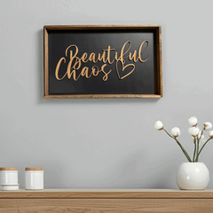 Framed wood sign with a black background and stained frame hanging on a grey wall above a wood table with white decorations