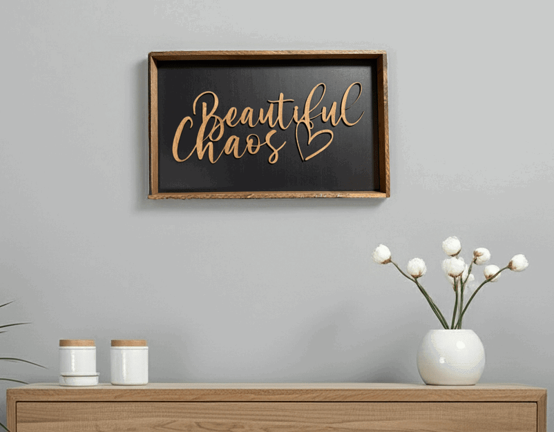 Framed wood sign with a black background and stained frame hanging on a grey wall above a wood table with white decorations