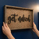 Wooden sign with 'get naked' text on a blue wall, hands holding the sign.