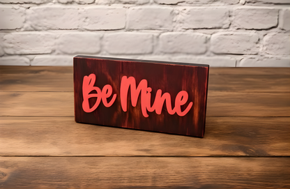 wood sign painted black with a faint red tint with the words Be Mine painted in red, sitting on a wood table with a white brick wall in the background