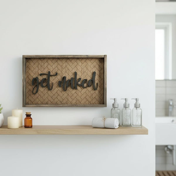 Wooden framed sign with laser engraved herringbone pattern and laser cut get naked hanging on a wall in a bathroom 
