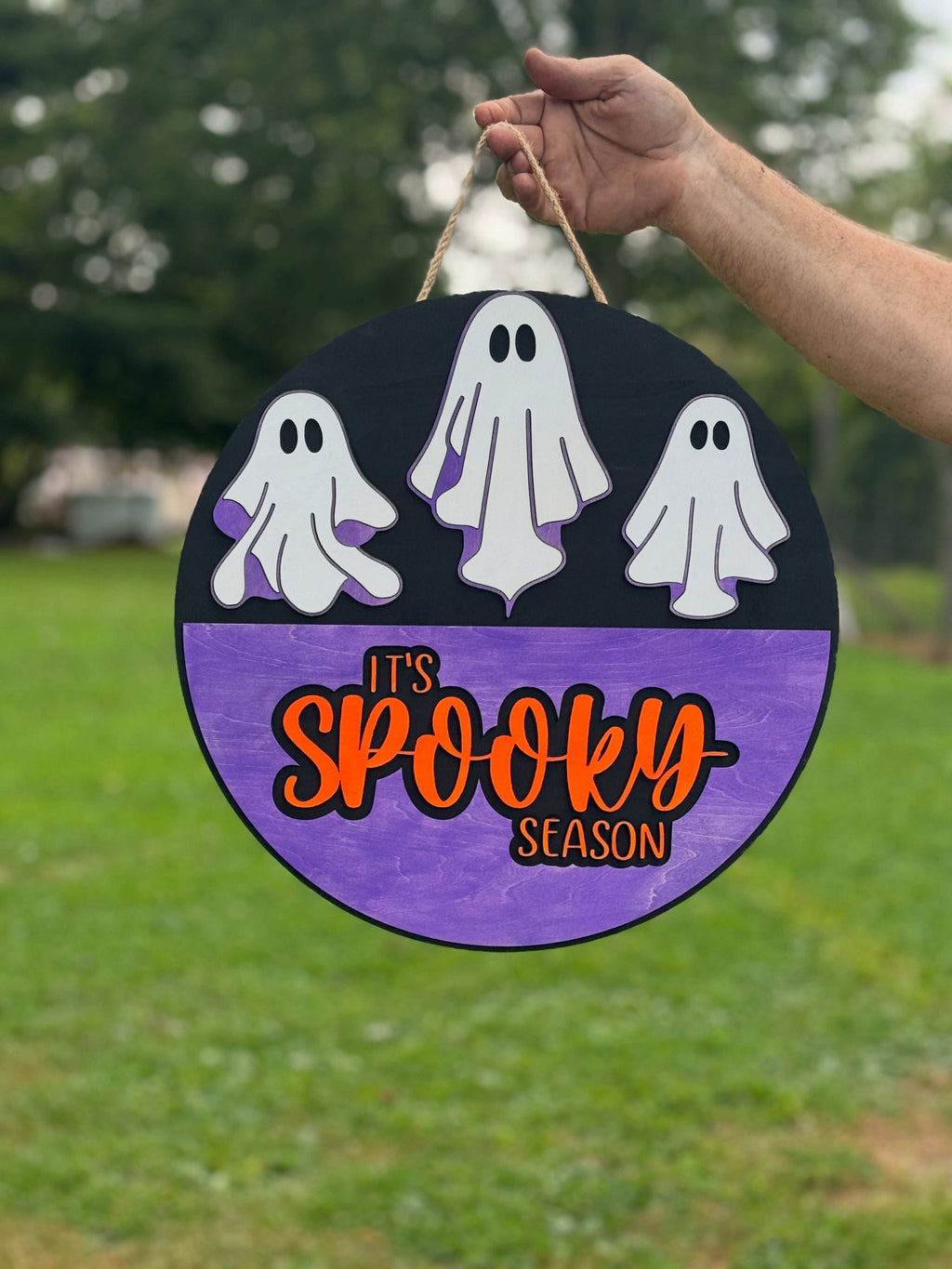 Decorative sign with ghosts and 'It's Spooky Season' text held outdoors.