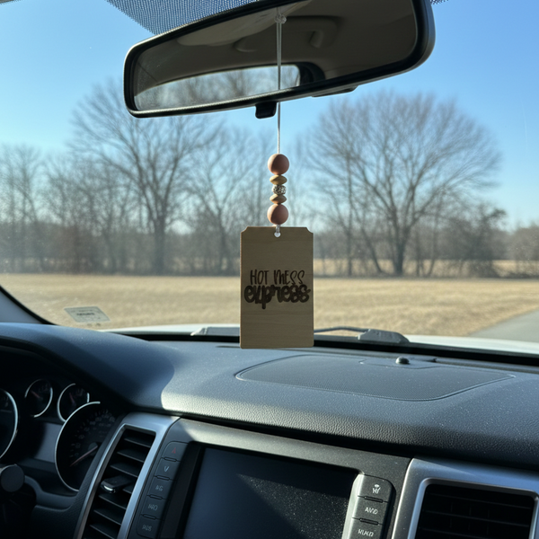 Wooden car freshener that says hot mess express with light pink beads hanging from a rearview mirror
