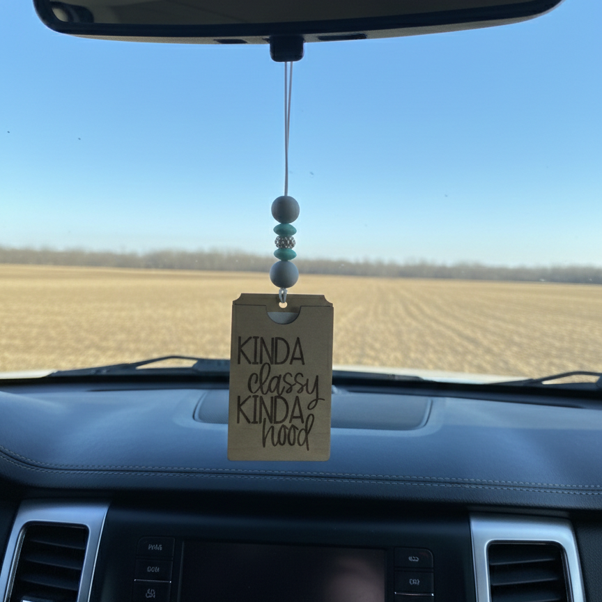 Wooden car freshener with the phrase engraved "kinda classy kinda hood" hanging from a rearview mirror