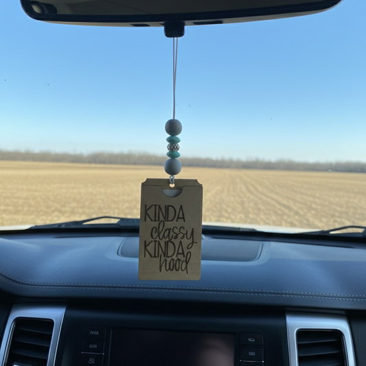 Wooden car freshener with the phrase engraved "kinda classy kinda hood" hanging from a rearview mirror