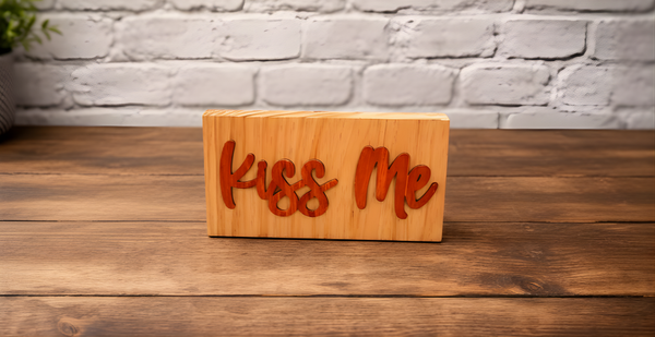 wood sign with the saying kiss me, sitting on a wood table with a white brick wall background