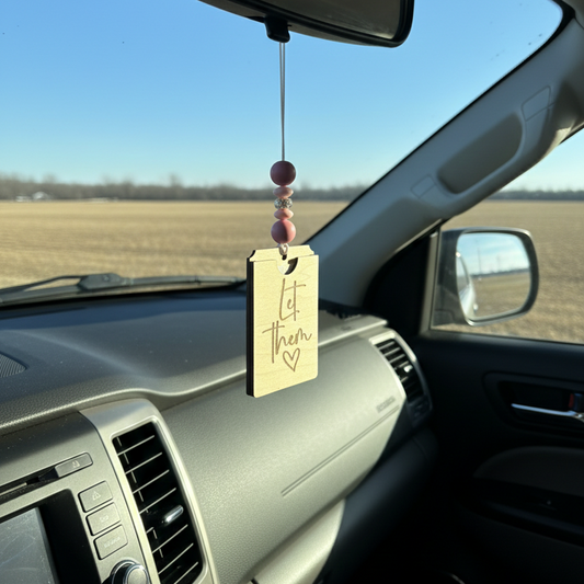 wooden car freshener hanging from a rearview mirror with the phrase engraved "let them"