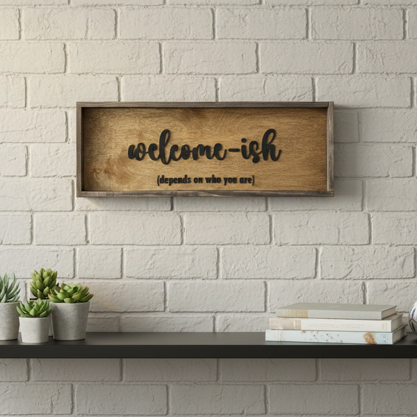 Wooden framed sign with 'welcome-ish' depends on who you are text on a white brick wall above a shelf with plants and books.