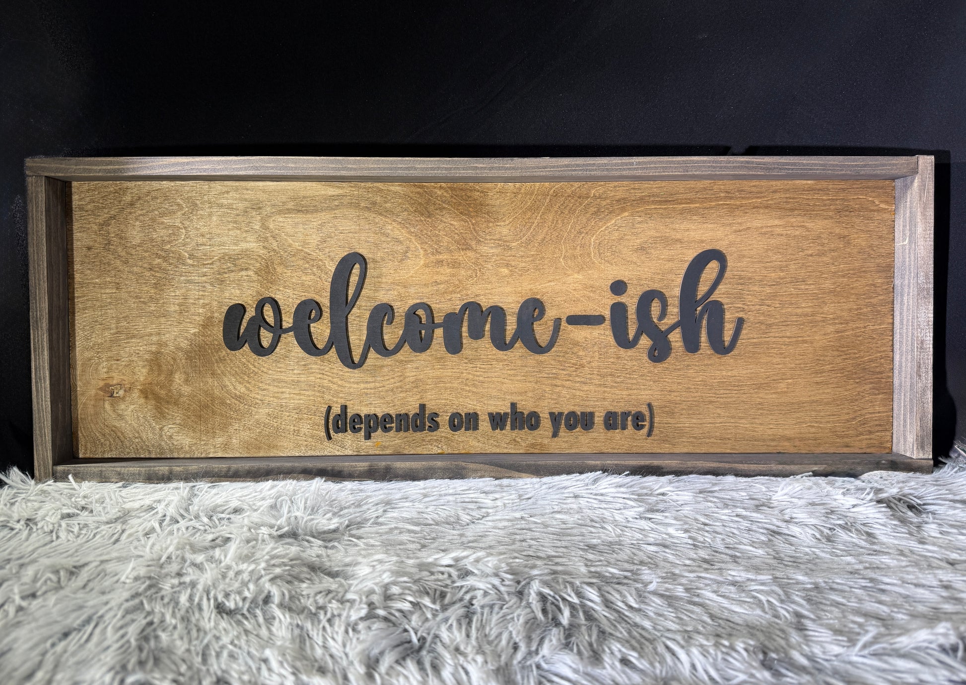 Wooden framed sign with 'welcome-ish (depends on who you are)' text on a textured surface