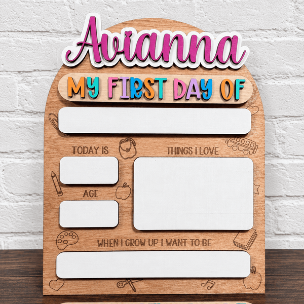 Personalized wooden first-day-of-school milestone sign with text reading 'Avianna' and 'MY FIRST DAY OF', plus labeled boxes for today's age, things I love, and when I grow up I want to be. The sign has colorful bubble-letter text and is shown against a white brick wall.
