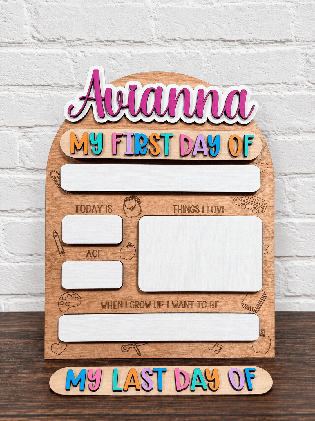 Personalized wooden first-day-of-school milestone sign with text reading 'Avianna' and 'MY FIRST DAY OF', plus labeled boxes for today's age, things I love, and when I grow up I want to be. The sign has colorful bubble-letter text and is shown against a white brick wall.