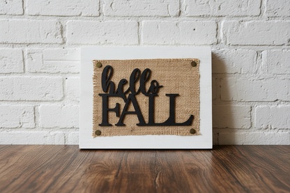 A square white-framed decorative sign leaning against a white brick wall on a dark wood surface. The sign features a burlap-textured center with black cutout lettering that reads 'hello FALL'.