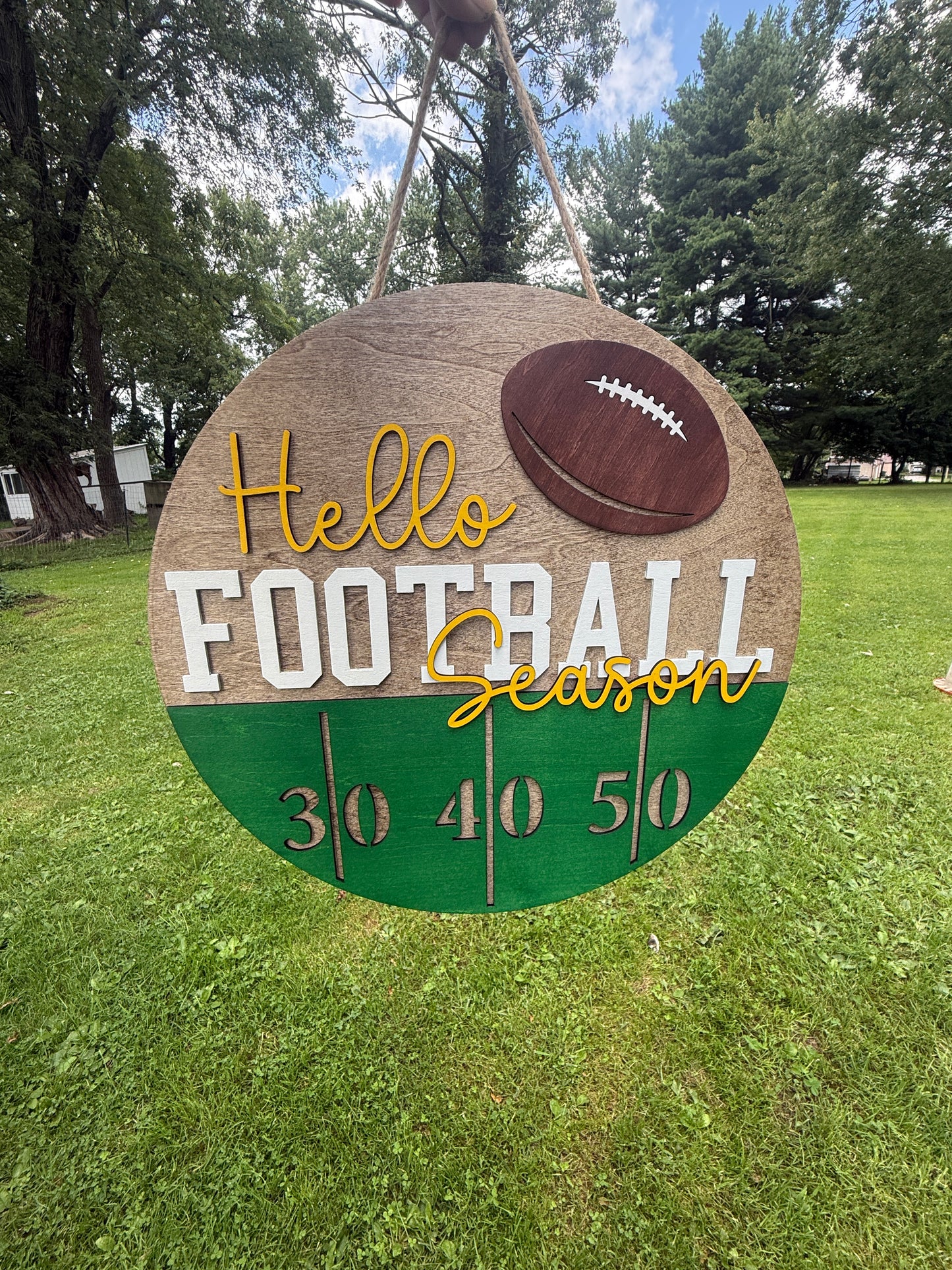 A round wooden hanging sign with a football theme, featuring the text 'Hello Football Season' and yard line numbers 30, 40, 50, set against a grassy outdoor background.