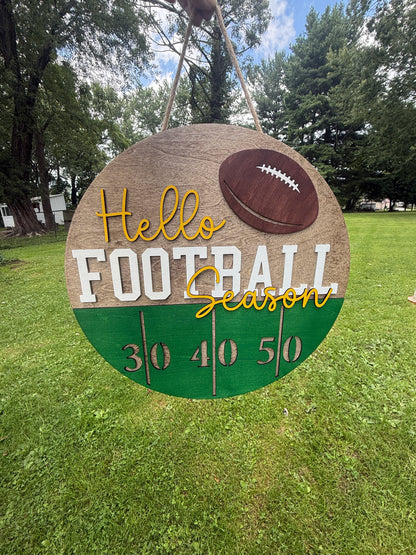 A round wooden hanging sign with a football theme, featuring the text 'Hello Football Season' and yard line numbers 30, 40, 50, set against a grassy outdoor background.