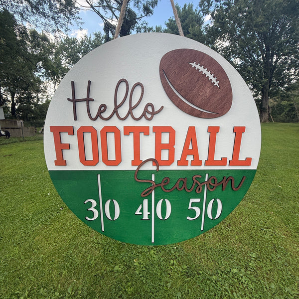 A round sign hanging from a rope, featuring the words 'Hello FOOTBALL Season' with a football graphic and a green scoring track at the bottom, photographed on grass with trees and sky behind it.