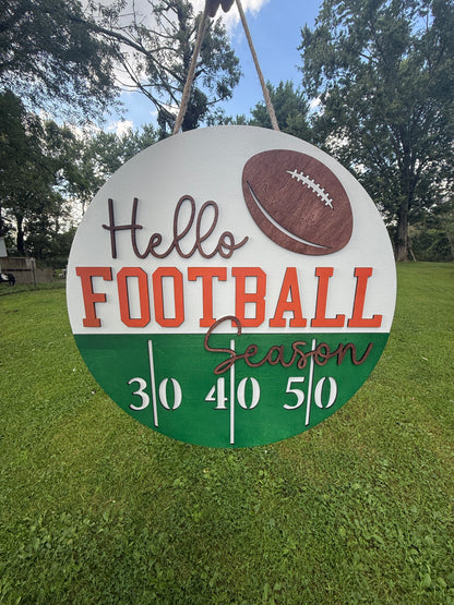 A round sign hanging from a rope, featuring the words 'Hello FOOTBALL Season' with a football graphic and a green scoring track at the bottom, photographed on grass with trees and sky behind it.