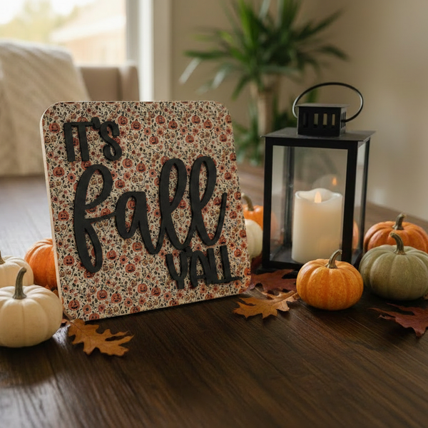A decorative wooden sign reading 'It's Fall Y'all' in black script over a muted floral and pumpkin pattern, styled on a dark wood table with small pumpkins, autumn leaves, a black lantern with a white candle, and a softly blurred indoor background.