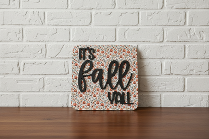 A square decorative sign with the phrase 'It's Fall VALL' in black lettering, featuring a small orange and beige floral pattern background, displayed on a wooden surface against a white brick wall.
