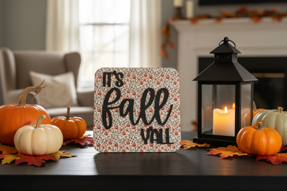 A square decorative sign with the phrase 'It's fall Y'all' in black script over a small floral and pumpkin pattern, styled on a dark table with pumpkins, autumn leaves, a black lantern with a lit candle, and a softly blurred living room background.
