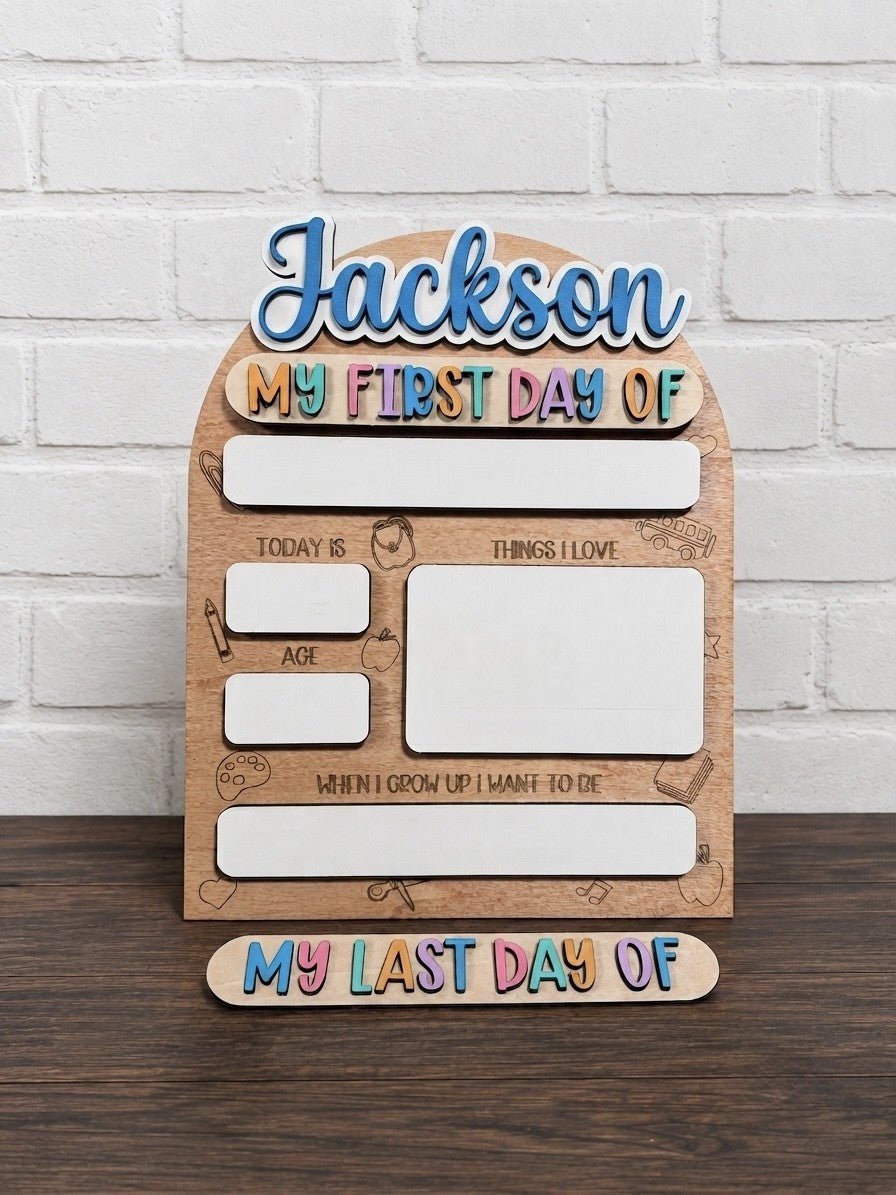 Personalized wooden first-day-of-school milestone board with the name 'Jackson' at the top, colorful text reading 'My First Day Of,' and a matching bottom piece reading 'My Last Day Of,' displayed on a dark wood surface against a white brick wall.