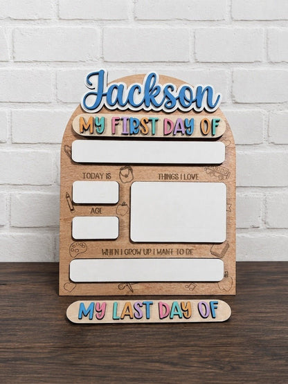Personalized wooden first-day-of-school milestone board with the name 'Jackson' at the top, colorful text reading 'My First Day Of,' and a matching bottom piece reading 'My Last Day Of,' displayed on a dark wood surface against a white brick wall.